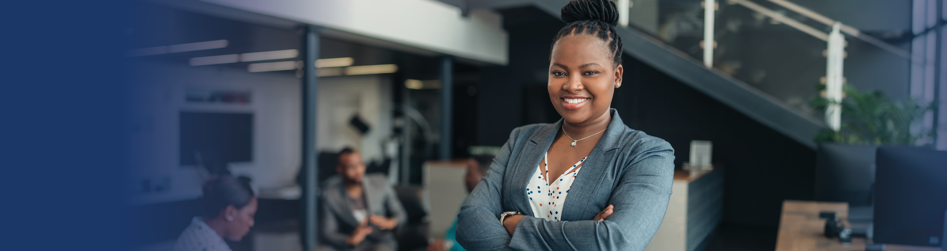 Professional woman stands with her arms crossed in a workplace setting with colleagues in the background.